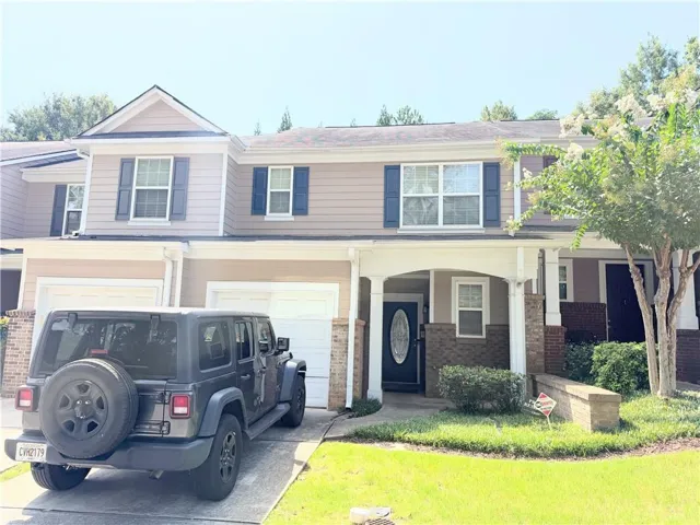 Two-story townhome with tan siding and dark shutters, featuring a covered entry porch supported by white columns, a single-car garage, and a manicured lawn