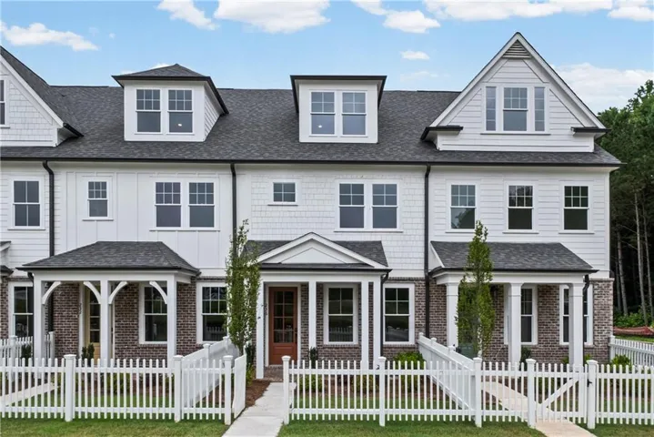 Modern farmhouse-style townhome featuring white siding, dark shingle roofing, brick accents, multiple dormer windows, and a white picket fence