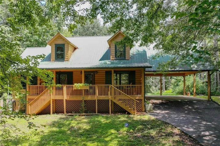 Log cabin exterior featuring a covered porch with wood railings and lattice, multiple dormer windows, a green metal roof, and an attached carport