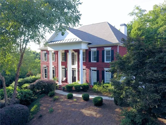 Red brick exterior with white trim, columned front porch, dark shutters, and a sloped, landscaped front yard