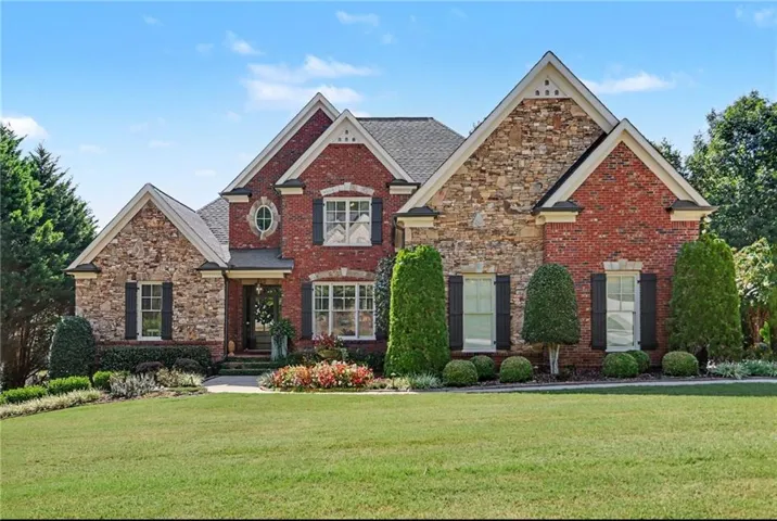 Brick and stone facade, gabled rooflines, black louvered shutters, decorative oval window, mature landscaping