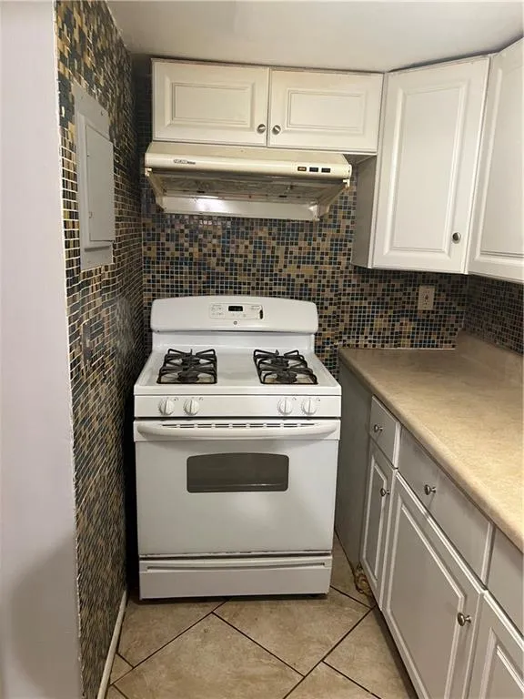 Kitchen featuring light tile floors, ventilation hood, white gas range oven, white cabinets, and backsplash