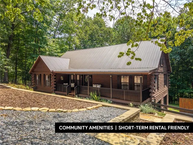 Log cabin exterior featuring a metal roof, expansive covered porch with dark railing, and a sloped, landscaped front yard with gravel and mulch