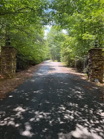Gated entrance featuring stone columns with lantern-style lighting and a paved driveway surrounded by mature trees