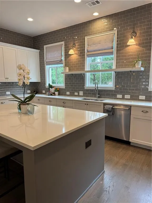 Kitchen featuring a substantial island with a white countertop, wood-finish flooring, white cabinetry, a full-height gray subway tile backsplash, and a stainless steel dishwasher