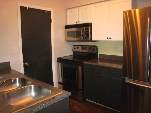 Kitchen featuring stainless steel appliances, dark lower cabinetry, white upper cabinetry, a light green tile backsplash, and a double basin stainless steel sink with a dark countertop