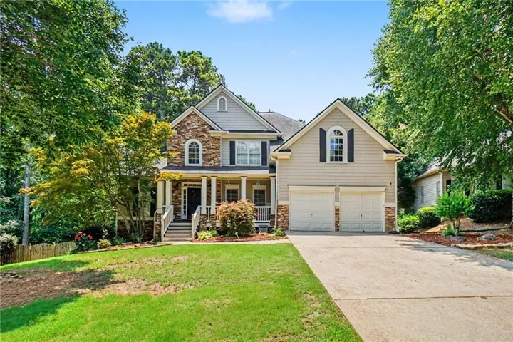 Traditional-style residence featuring a stone and siding facade, a covered front porch with white columns and railings, a two-car attached garage, a paved driveway, and mature landscaping