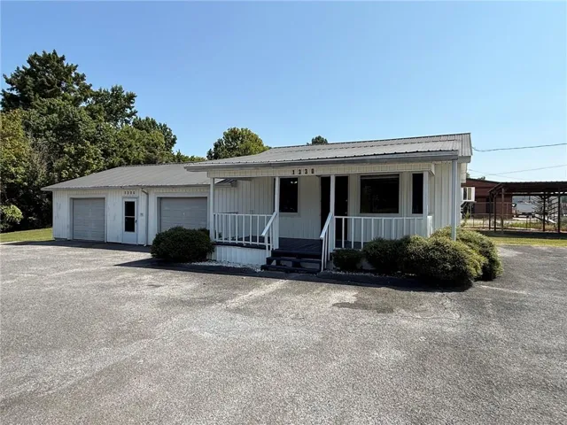 White exterior siding, gray metal roof, covered front porch with white railing, paved parking area, and an attached two-car garage