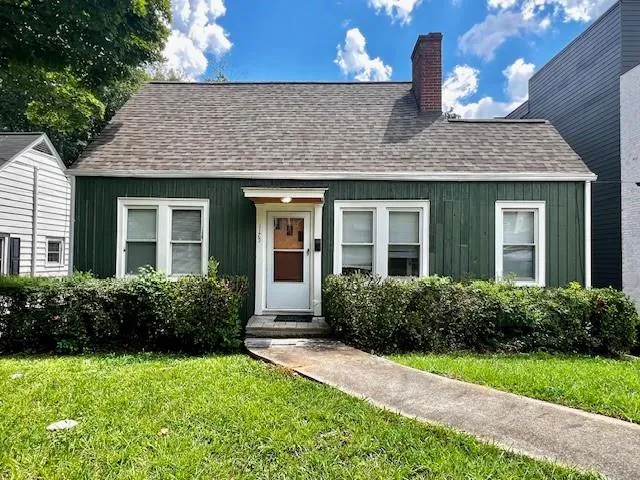 Exterior featuring dark green vertical siding, white trim windows, a white entry door, an asphalt shingle roof, and a red brick chimney