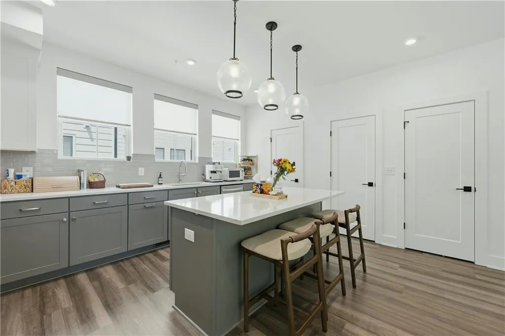 Kitchen featuring an eat-at island with a quartz countertop, wood-finish flooring, shaker-style cabinetry, a tile backsplash, and three pendant light fixtures