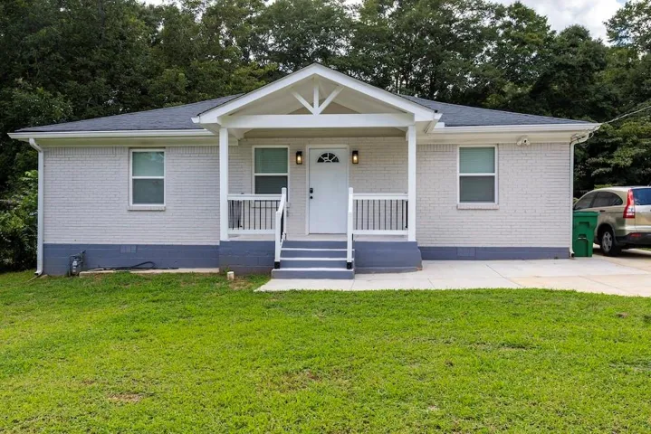 White brick exterior with a covered porch featuring white railings, dark gray foundation, and a dark shingle roof
