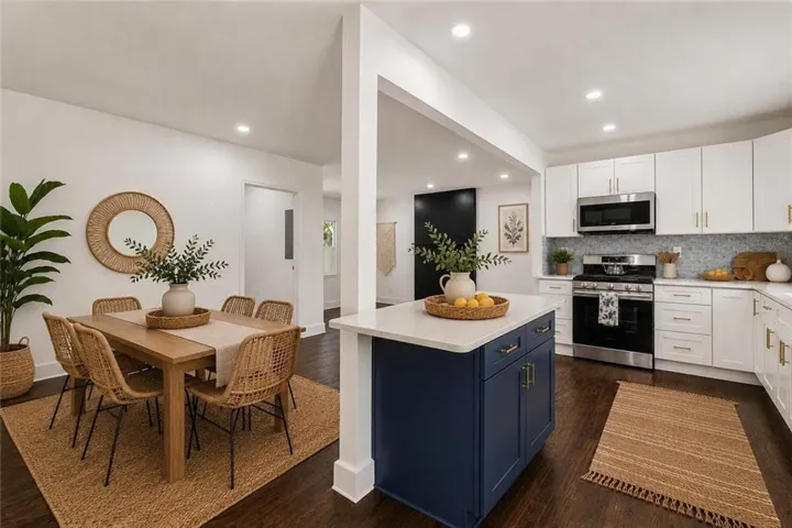 Open-concept kitchen and dining area featuring dark wood-finish flooring, white cabinetry with brass-finish hardware, a blue kitchen island with a white countertop, and recessed lighting