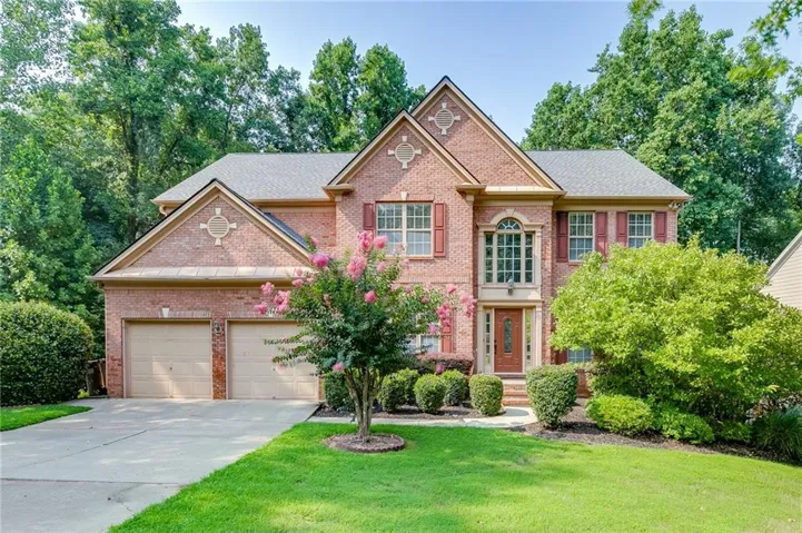 Traditional brick exterior featuring a two-car garage, manicured lawn, established landscaping, and decorative red shutters
