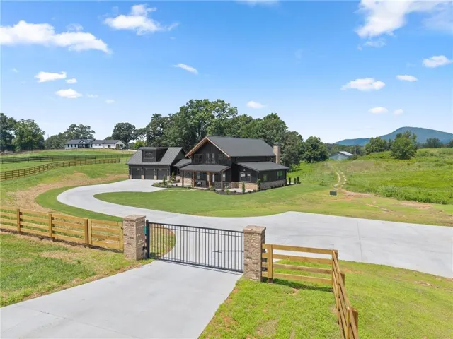 Modern farmhouse residence set on expansive grounds featuring a curved concrete driveway, black metal entry gate with wooden posts, manicured lawn, wooden ranch-style fencing, mature tree line, and distant mountain backdrop
