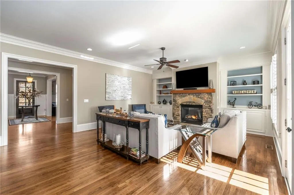 Living room featuring built in shelves, dark wood-type flooring, ornamental molding, a ceiling fan, and a stone fireplace