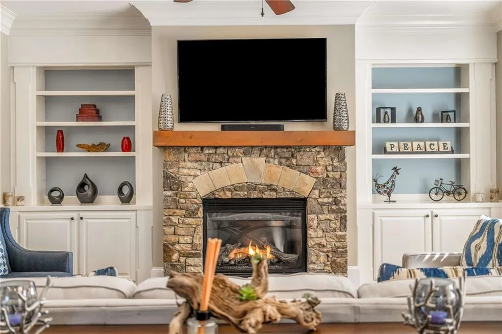Living area with ornamental molding, a stone fireplace, built in features, and a ceiling fan