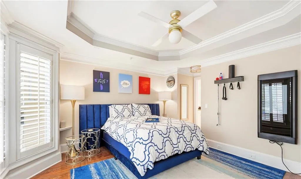 Tray ceiling with crown molding, bay window with plantation shutters, wood-finish flooring, and a central ceiling fan