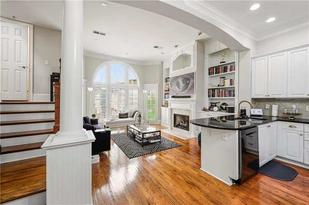 Open-concept interior featuring wood-finish flooring, a kitchen island with stone countertops, a fireplace with custom built-in cabinetry, an arched window with shutter treatments, and a staircase with wood treads