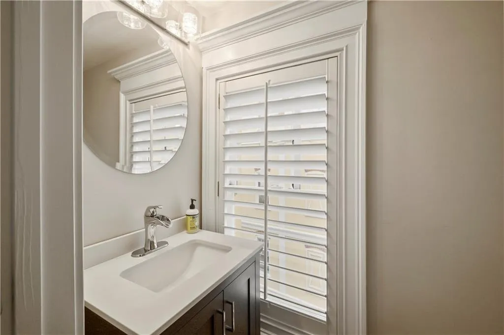 Vanity featuring a stone countertop, integrated basin, chrome faucet, circular mirror, and window with white plantation shutters