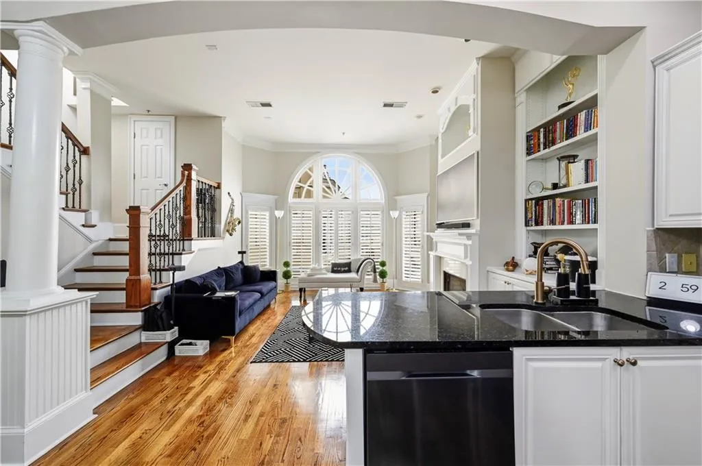 Wood-finish flooring, arched window with decorative mullions, built-in floor-to-ceiling cabinetry, staircase with decorative iron balusters, and kitchen island with dark stone countertop