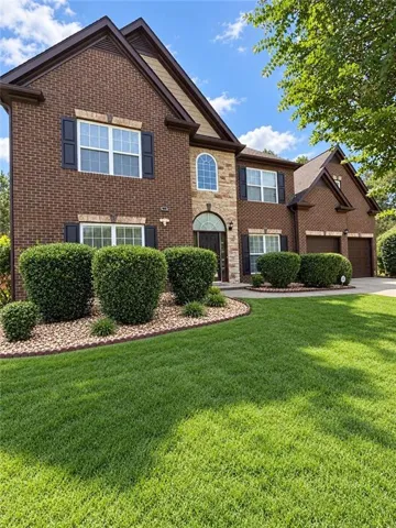 Brick and stone facade, arched entryway, gabled rooflines, dark shutters, and a manicured lawn with structured landscaping