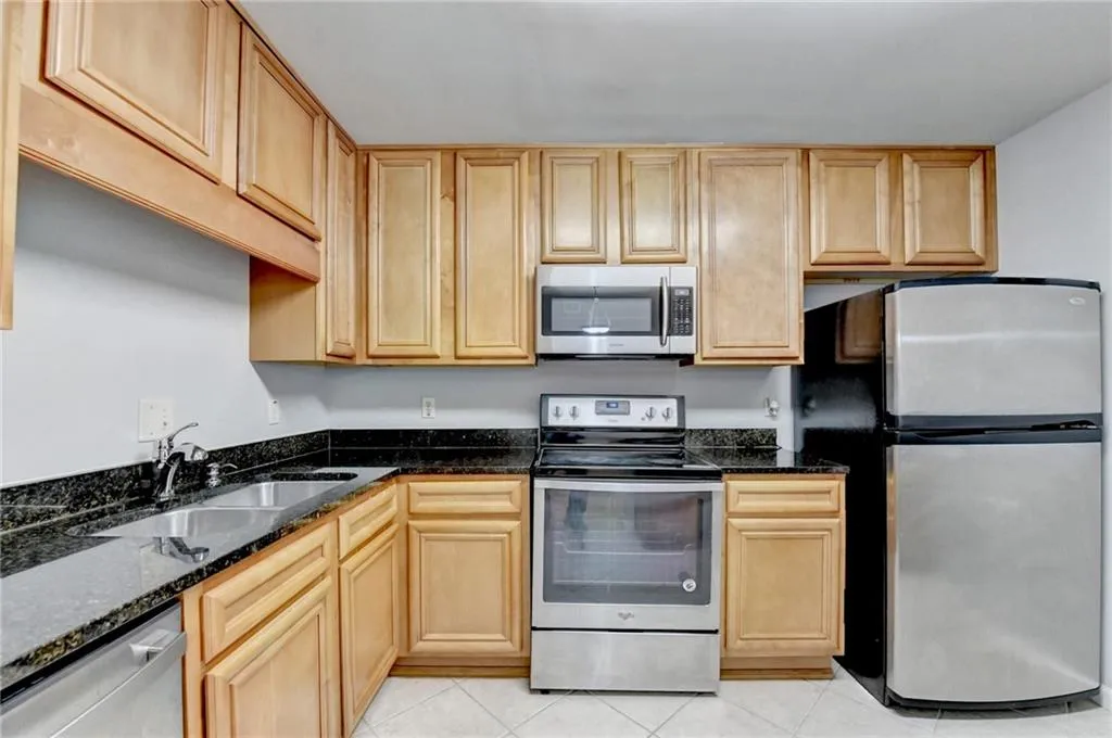 Kitchen with stainless steel appliances, dark stone countertops, light wood finish cabinets, and light tile patterned floors