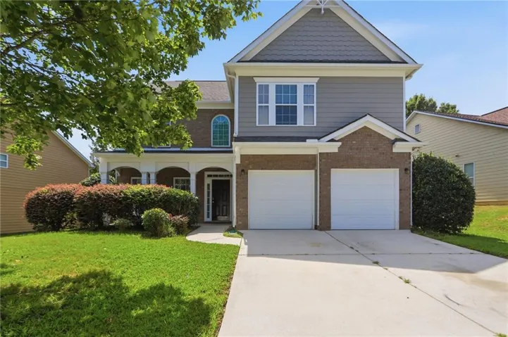 Two-car garage with white panel doors, brick and siding exterior, front-load covered entry, manicured lawn with mature landscaping, and two-story colonial-style architecture with gabled roofline