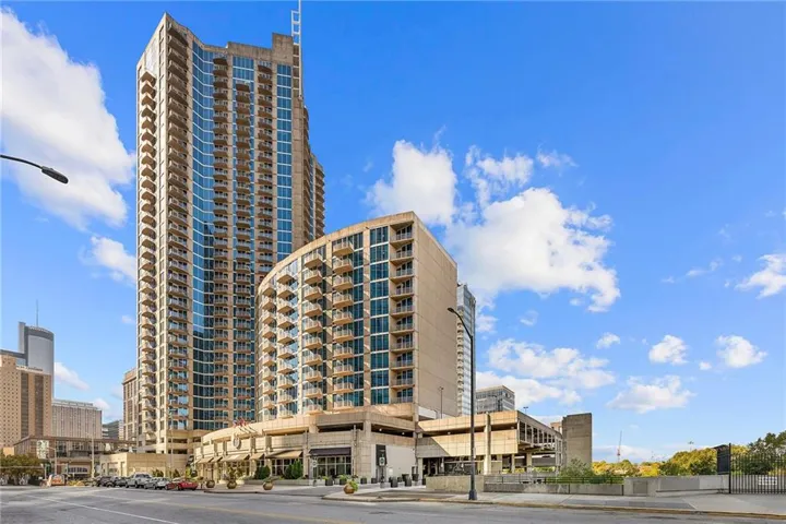 Contemporary high-rise building featuring numerous private balconies, a curved facade, a ground-level retail presence, and multi-level parking structures