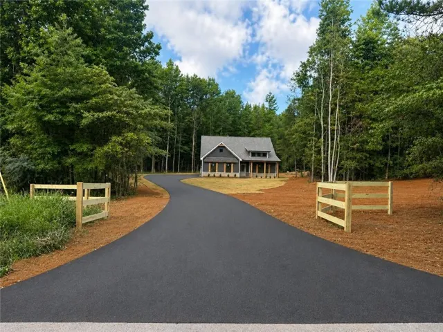Paved asphalt driveway with a natural wood fence, surrounded by mature trees and a newly constructed home with a covered front porch and stone accents