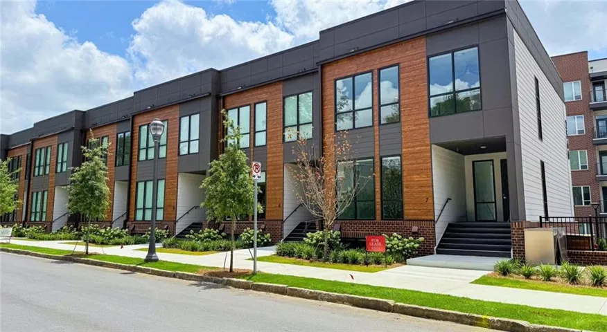 Modern townhome exterior featuring multi-level windows, a mixed material facade with wood paneling and dark siding, a covered entry, and a paved walkway with planted landscaping
