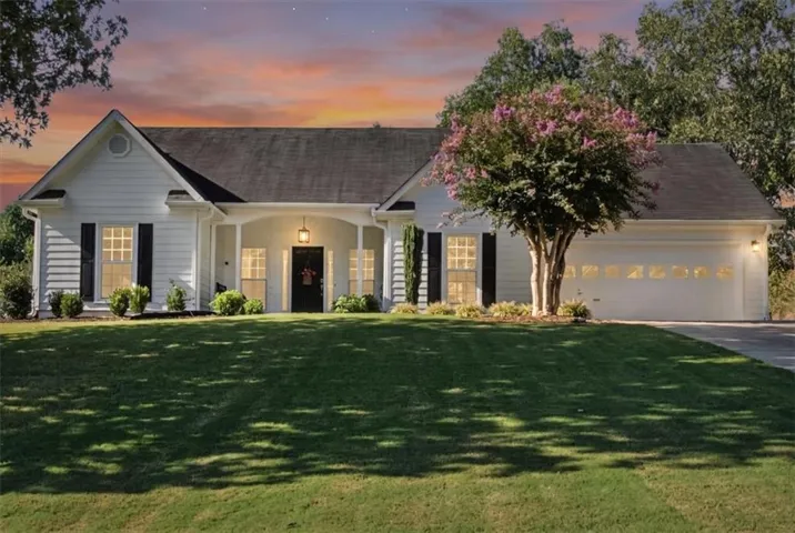 White-sided exterior with dark shutters, covered front entry, attached two-car garage, manicured lawn, and mature tree