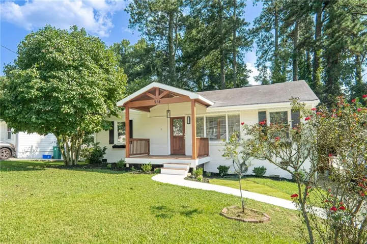 White exterior with dark shutters, covered front porch with wood-finish columns and railing, wood-finish front door, and established landscaping
