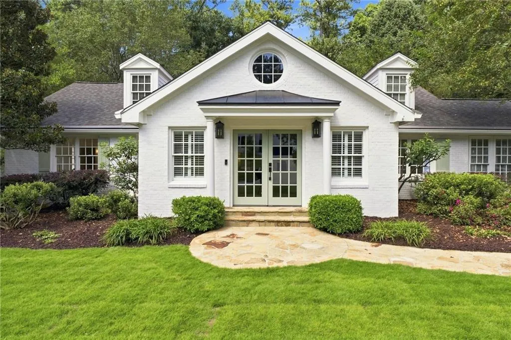 White brick exterior with a dark-pitched roof, featuring a portico entry with light-colored French doors, flanked by symmetrical windows with white grilles