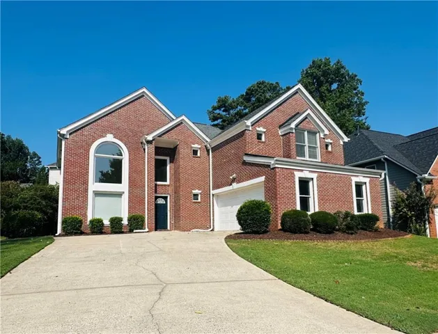Brick exterior with white trim, arched window feature, attached garage, concrete driveway, and established shrubbery