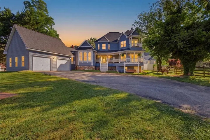Victorian-inspired residence featuring a two-car garage, expansive wrap-around porch, stone foundation accents, multiple gables, and a mature tree-lined property boundary