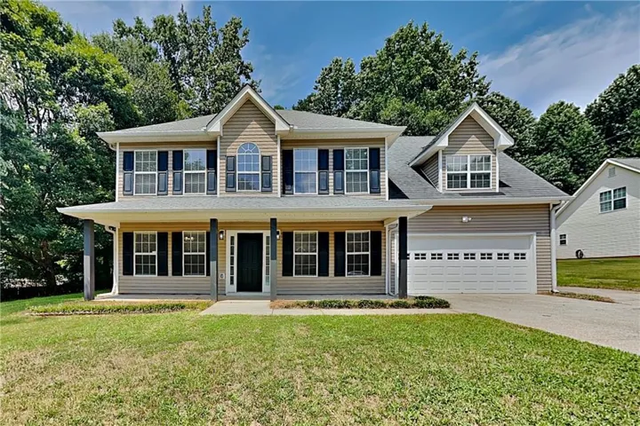Two-story exterior with light beige siding and dark shutters, featuring a covered front porch with four columns, an attached two-car garage with a panel door, and a front yard with established landscaping