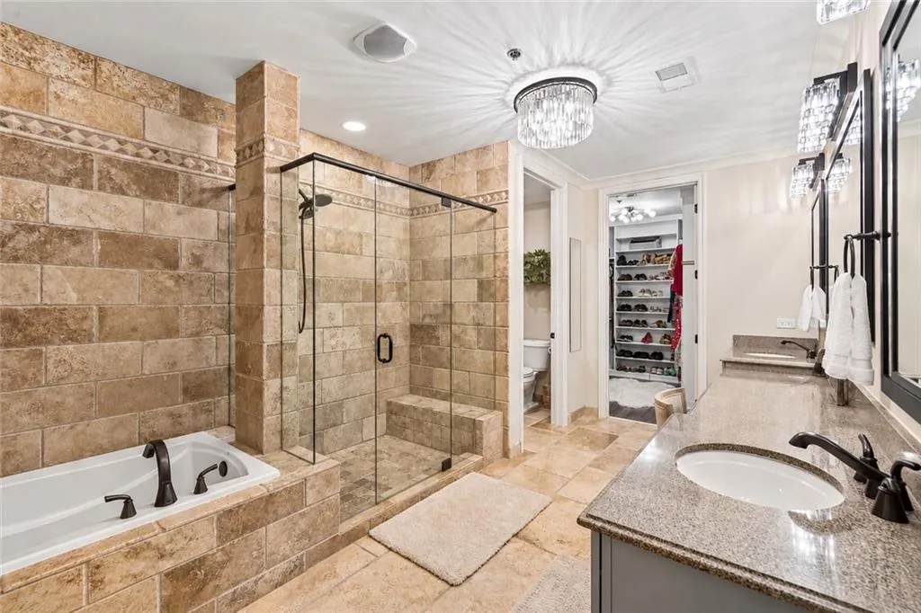 Tiled bathroom featuring a built-in bathtub, a walk-in shower with a glass enclosure, a dual vanity with a stone countertop, and a crystal chandelier