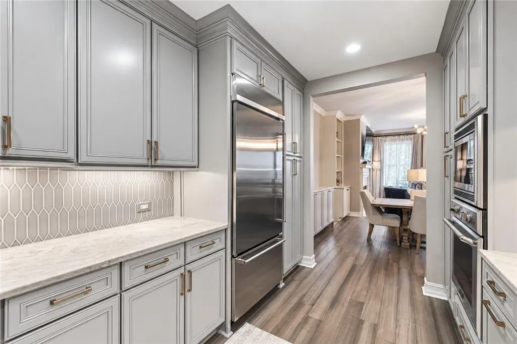 Light gray cabinetry with gold-tone hardware, stainless steel refrigerator, light gray geometric tile backsplash, light wood-finish flooring, and recessed lighting