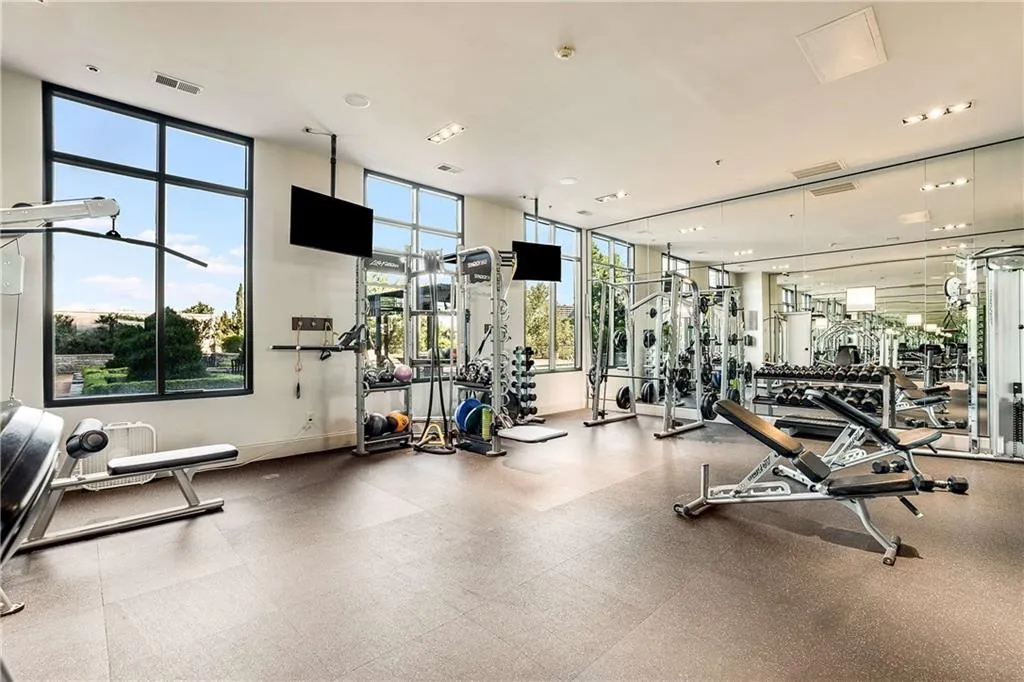 Expansive fitness center featuring resilient rubberized flooring, abundant natural light from multiple windows, recessed ceiling lighting, wall-mounted mirrored panels, and integrated televisions