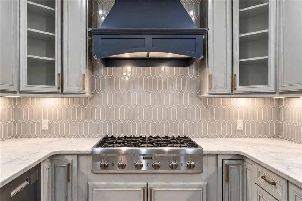 Stainless steel gas range with a custom range hood, marble-finish countertops, geometric tile backsplash, and light-toned cabinetry with glass-front uppers