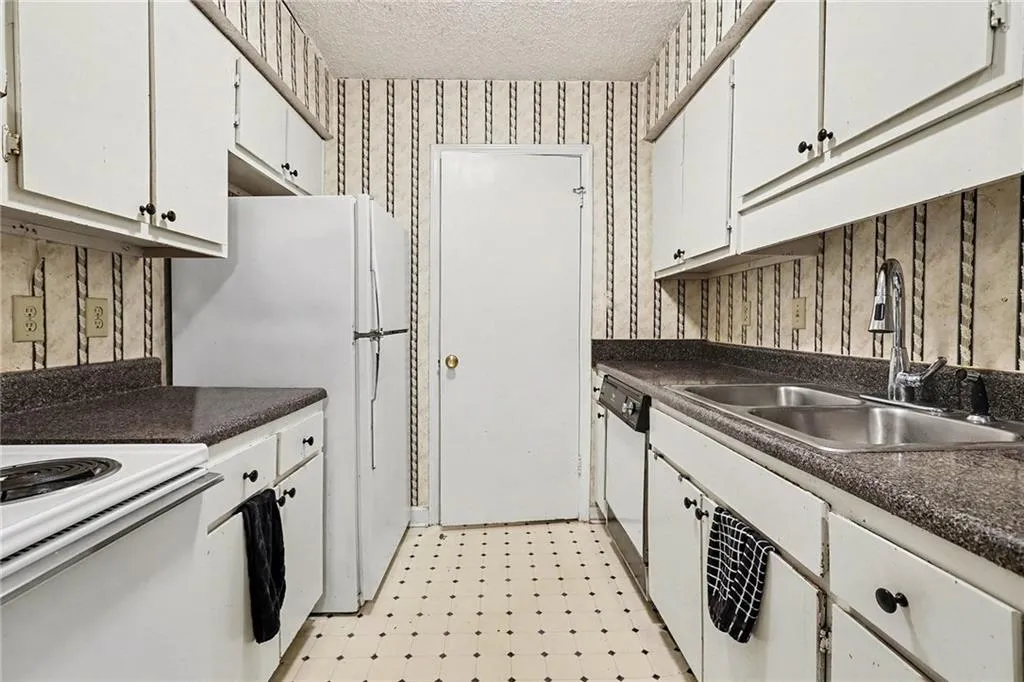 Kitchen featuring light floors, dark countertops, white appliances, wallpapered walls, and a textured ceiling
