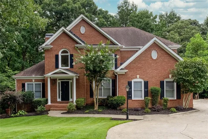 Traditional brick exterior residence featuring a prominent gabled roofline, arched windows, decorative shutters, a covered entry with white columns, and mature landscaping