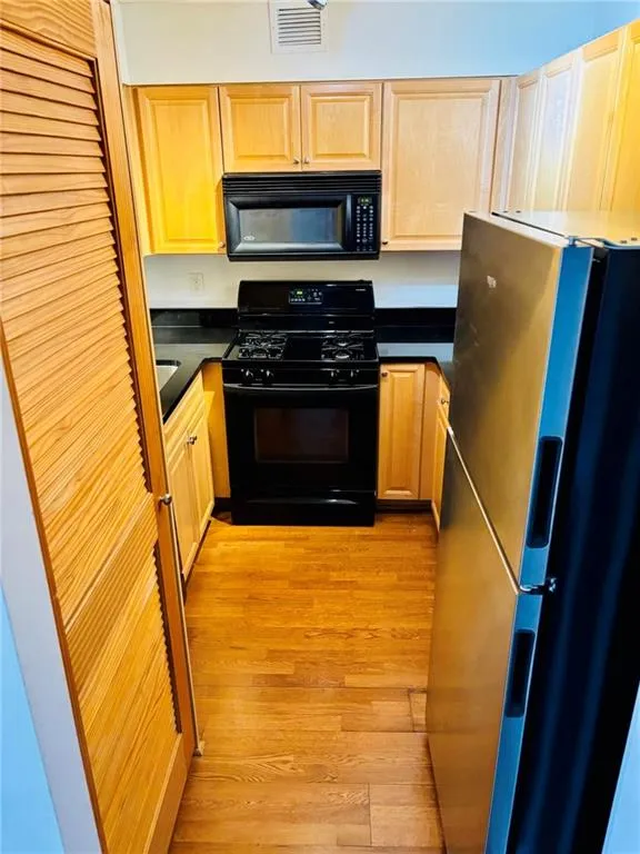 Kitchen with black appliances, light wood-type flooring, light brown cabinets, and dark countertops
