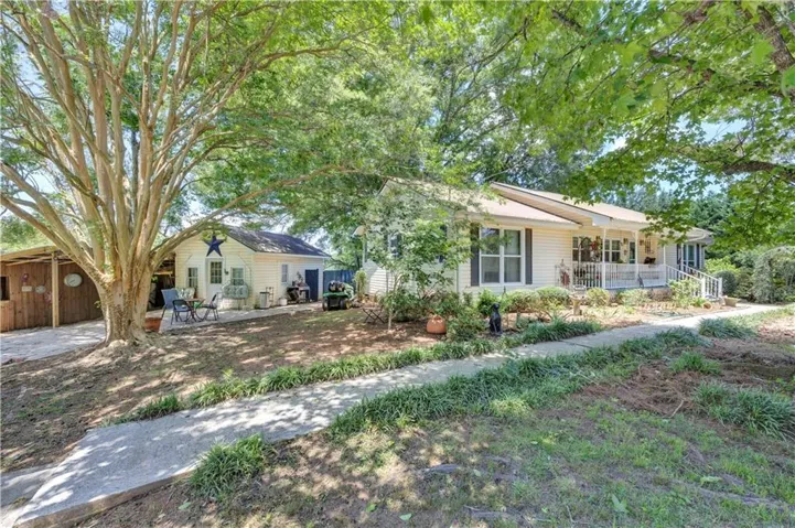 Cottage-style residence featuring a covered front porch with white railings, a concrete walkway, and mature trees providing natural canopy