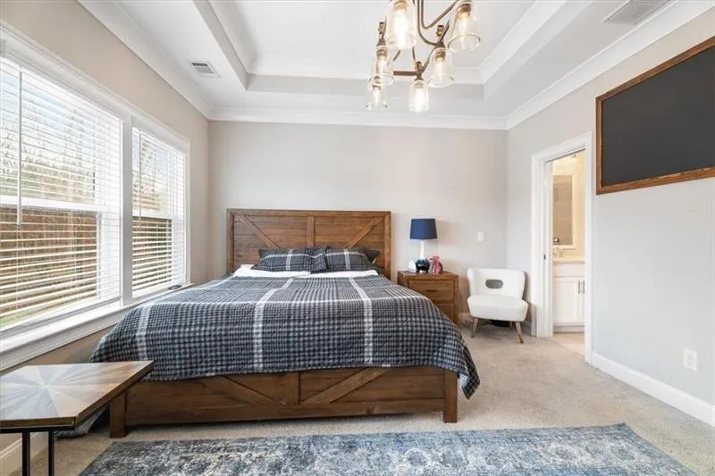 Bedroom featuring connected bathroom, a raised ceiling, light colored carpet, a chandelier, and ornamental molding
