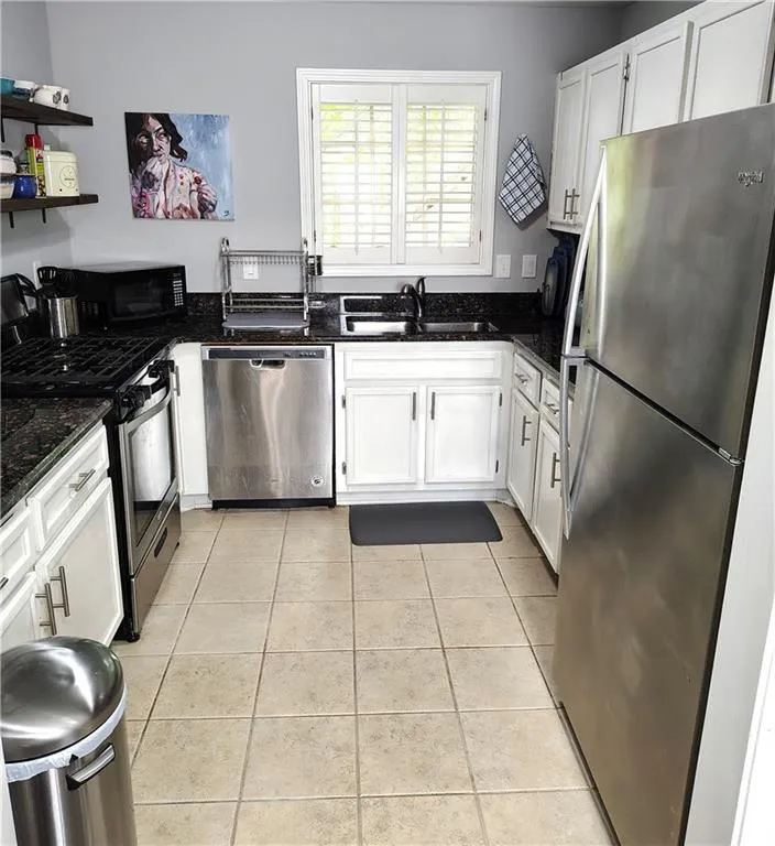 Kitchen with stainless steel appliances, dark stone countertops, a sink, light tile patterned flooring, and white cabinetry