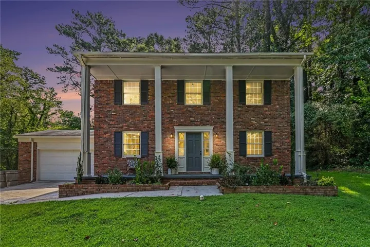Brick exterior with contrasting shutters, columned portico, attached garage, and mature perimeter landscaping