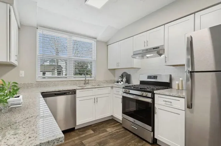 Kitchen featuring appliances with stainless steel finishes, under cabinet range hood, white cabinets, dark wood-style floors, and light stone counters