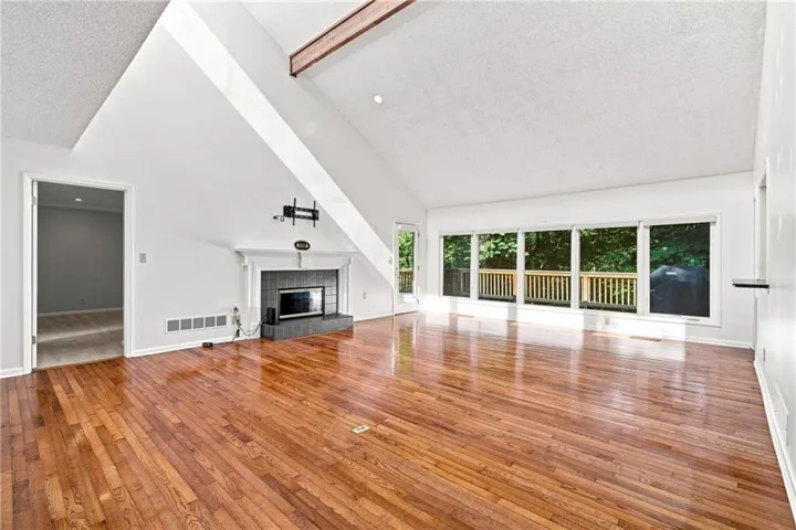 Vaulted ceilings with exposed wood beam, wood-finish flooring, fireplace with tile surround and mantel, and a wall of windows overlooking a deck