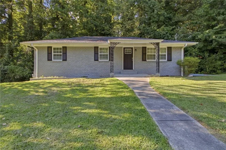 Grey brick exterior with a dark-tone entry door, covered porch with decorative metal columns, multiple windows with dark shutters, and a concrete walk